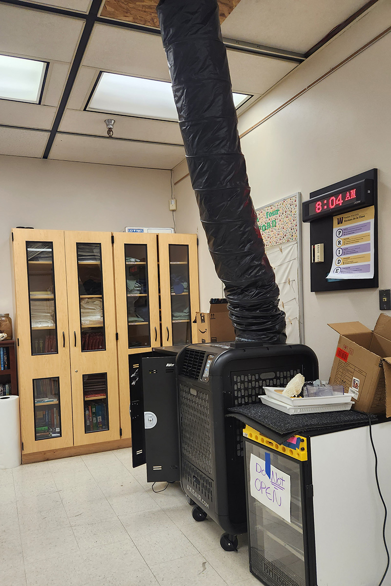 A portable air conditioner with a black plastic duct hose running up through a ceiling tile in an active WHS classroom, with a clock reading 8:04 AM
