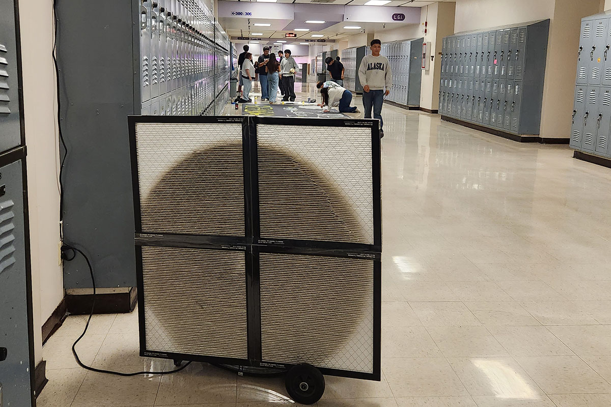 A makeshift air filtration unit built from four HVAC filters duct-taped together in front of a fan, parked in a WHS student hallway with students walking past in the background — the dark circular center shows heavy smoke contamination