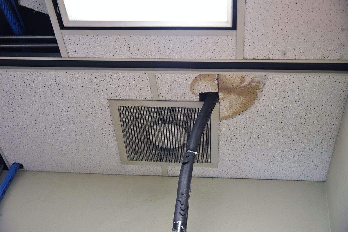 A WHS classroom ceiling tile with a large water and rust stain next to a failing HVAC vent, with a hose propped in as a makeshift repair