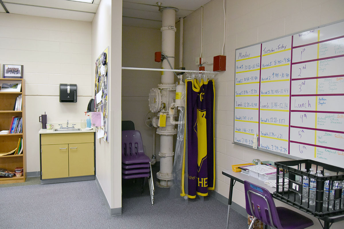 A WHS classroom with a shower curtain rod installed to conceal large exposed plumbing pipes running through the room, with student desks and a schedule board visible