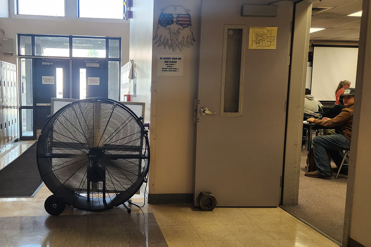 A large commercial barrel fan on wheels parked in a WHS hallway directly outside an active shop class, with students visible working at desks inside the classroom through the door window